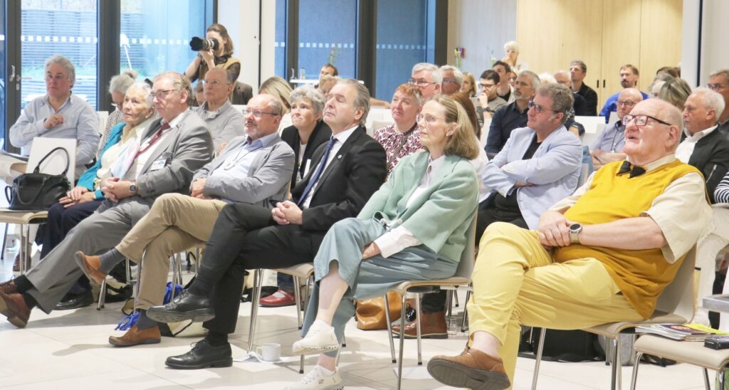 Menschen sitzen in einem hellen Raum auf Stühlen und blicken aufmerksam nach vorne. Die meisten tragen formelle oder gepflegte Kleidung, darunter Sakkos, Hemden, Blusen und Pullover. Im Hintergrund stehen weitere Personen, einige mit verschränkten Armen, andere mit Kameras. Die Atmosphäre wirkt konzentriert und ruhig.