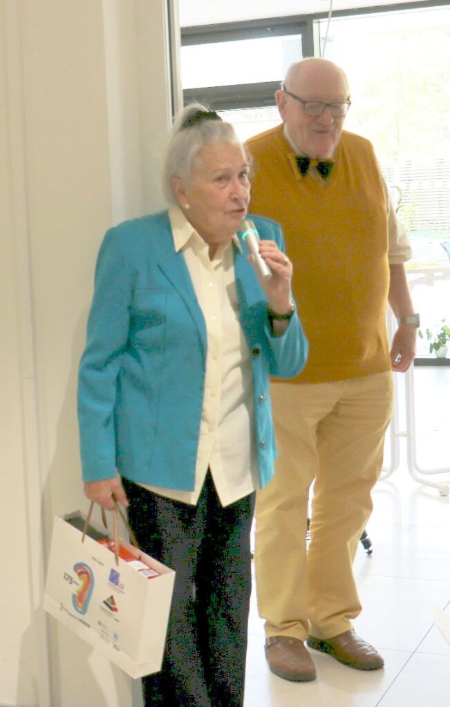 Ältere Frau mit grauem Dutt und blauem Blazer hält eine weiße Papiertüte und ein Mikrofon, hinter ihr steht ein älterer Mann mit Brille, gelbem Pullover und Fliege in einem hellen Raum.