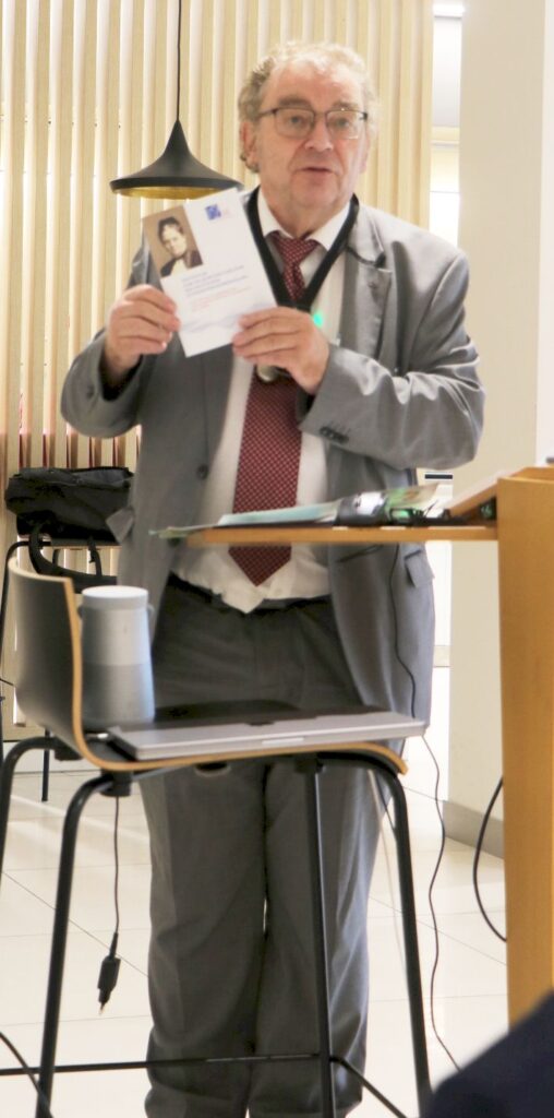 Älterer Mann mit grauem Anzug, roter Krawatte und Brille steht vor einem Pult und hält ein Buch mit einem Porträt auf dem Cover hoch.