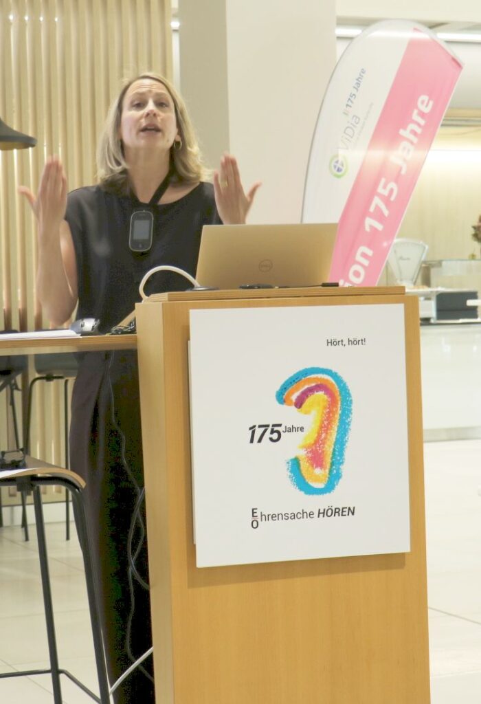 Frau steht hinter einem Rednerpult mit einem Plakat, das ein buntes Ohr und den Text '175 Jahre Ehrensache HÖREN' zeigt, sie gestikuliert mit erhobenen Händen.