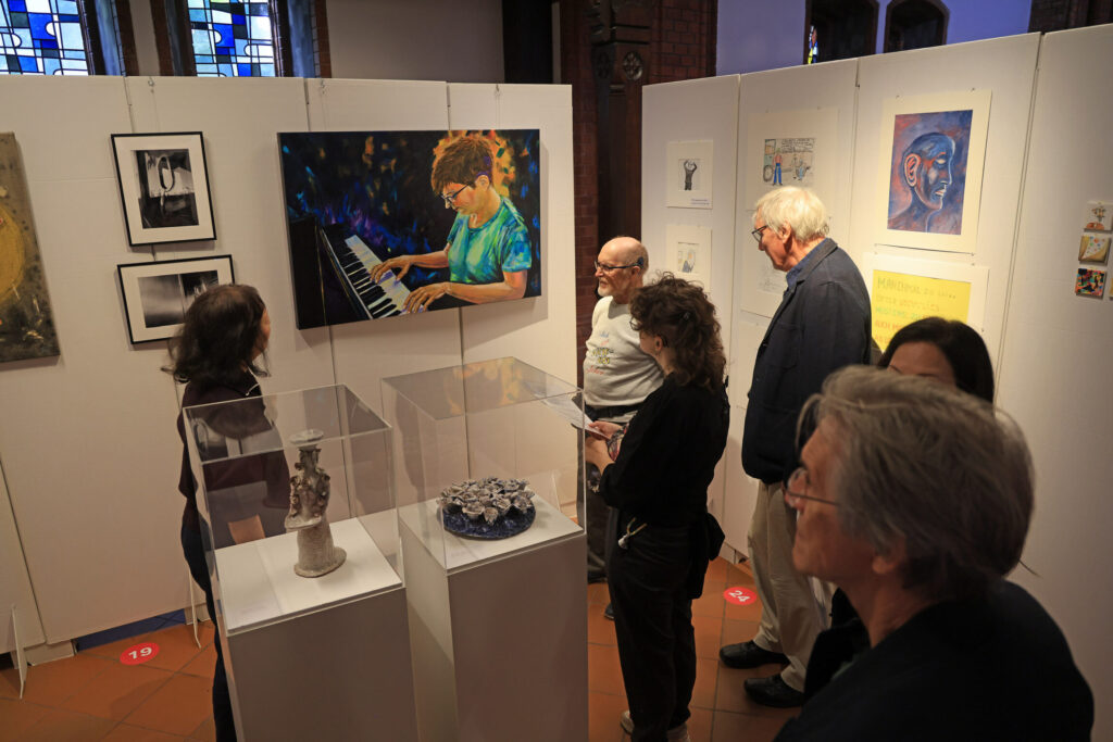 Mehrere Personen stehen in einem Ausstellungsraum und betrachten Kunstwerke, die an weißen Stellwänden hängen. Im Mittelpunkt hängt ein farbiges Gemälde einer Person am Klavier. Um das Bild herum sind weitere Kunstwerke, darunter Zeichnungen und Gemälde, zu sehen. Auf Podesten in Glasvitrinen stehen Skulpturen. Im Hintergrund sind bunte Glasfenster und ein Teil einer Backsteinwand sichtbar. Die Besucherinnen und Besucher tragen überwiegend dunkle Kleidung und betrachten die Exponate aus nächster Nähe.