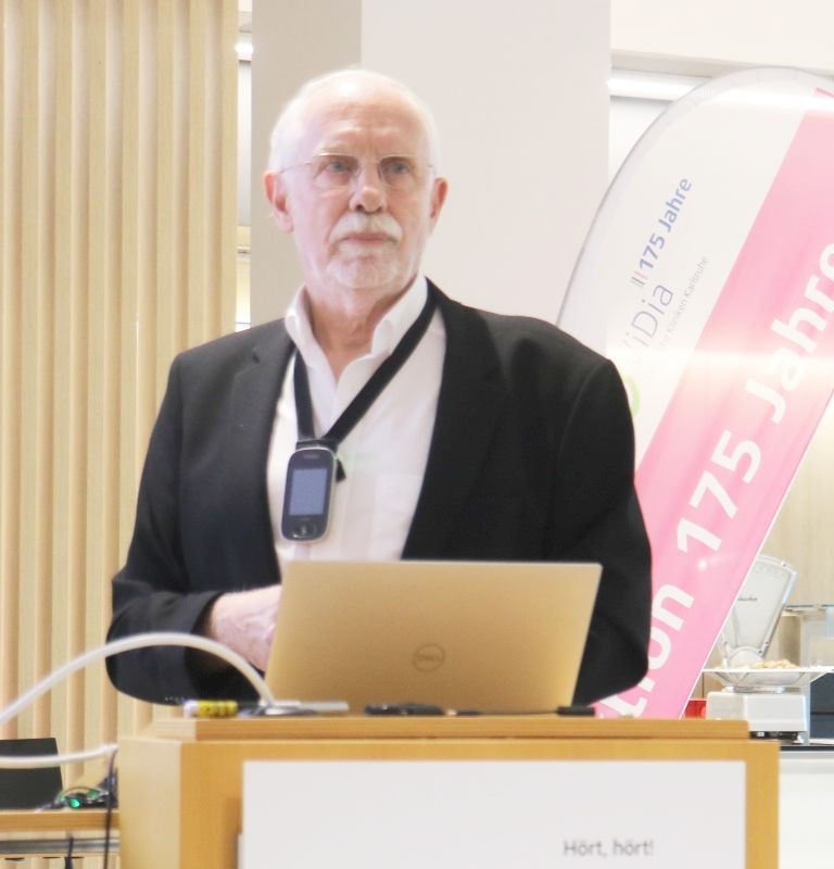 Älterer Mann mit weißem Haar und Bart steht hinter einem Rednerpult mit Laptop und trägt ein Mirkrofon um den Hals.