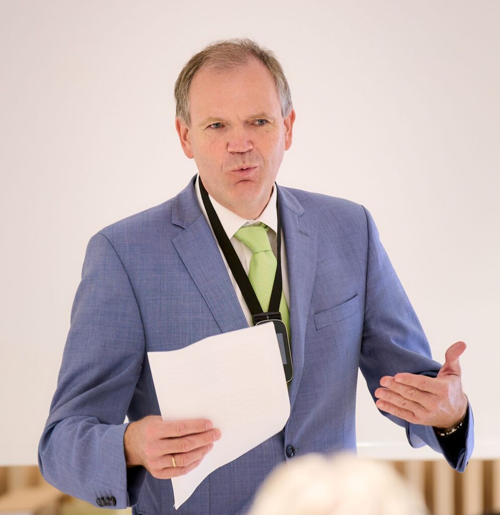 Mann im blauen Anzug mit grüner Krawatte hält ein Blatt Papier und gestikuliert während einer Rede oder Präsentation.