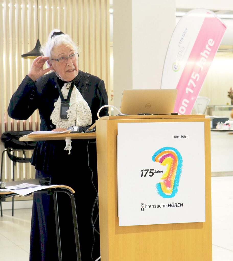 Ältere Frau mit weißem Haarknoten und schwarzer Kleidung steht an einem Rednerpult mit Laptop und hält sich eine Hand ans Ohr.