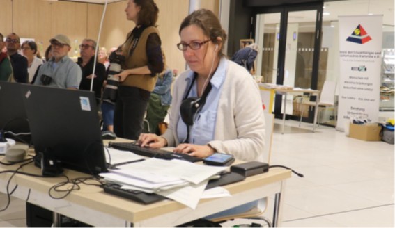 Frau mit Brille und weißer Jacke sitzt an einem Tisch mit Laptop, Tastatur und Papieren in einem hellen Raum mit mehreren Personen im Hintergrund.